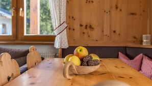 A wooden table with a bowl of fruit and pine cones, a window with curtains, and a checkered cushion.