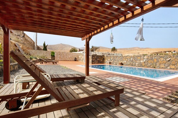 A wooden deck with lounge chairs, a swimming pool, and a stone wall.