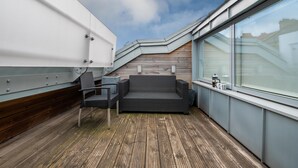 Um terraço no telhado com deck de madeira, um sofá de vime preto, uma cadeira de metal e uma grande janela com vista para o horizonte da cidade.
