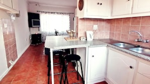 A compact kitchen with white cabinets, a granite countertop, and a sink. There are two black stools and a small table with a decorative item. - Ambiente Budget Accommodation (Upington)