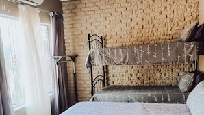 A room with a brick wall, a bed with a patterned bedspread, and a window with curtains. - Ambiente Budget Accommodation (Upington)