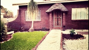 A brick house with a thatched roof, a small front yard with a lawn, a gravel pathway, and a potted plant. - Ambiente Budget Accommodation (Upington)