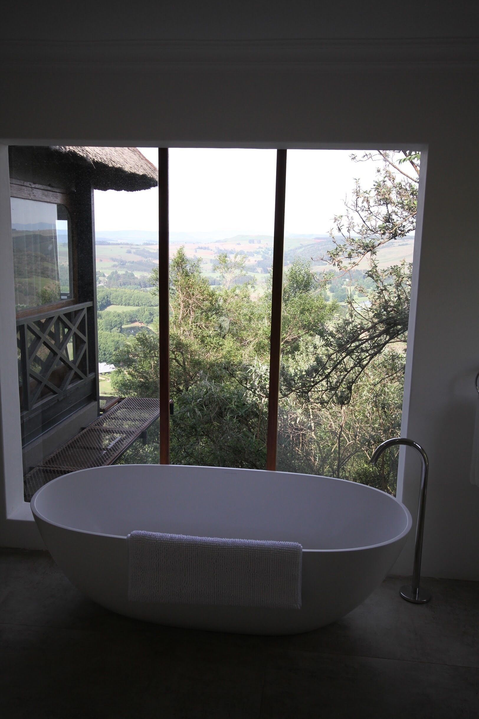 Un baño moderno con una bañera independiente, vistas a un paisaje exuberante y un balcón con techo de paja.