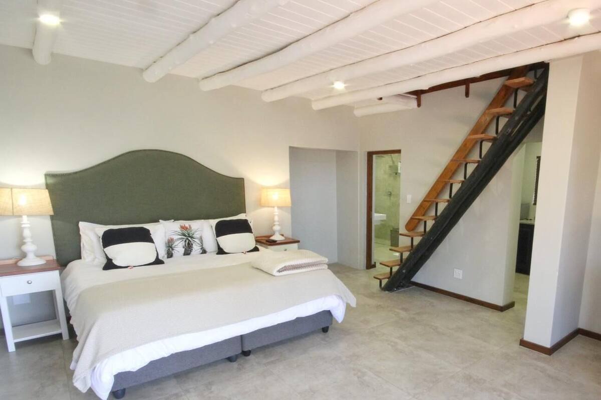 A bedroom with a large bed, a nightstand, a lamp, a staircase, and a door leading to another room.