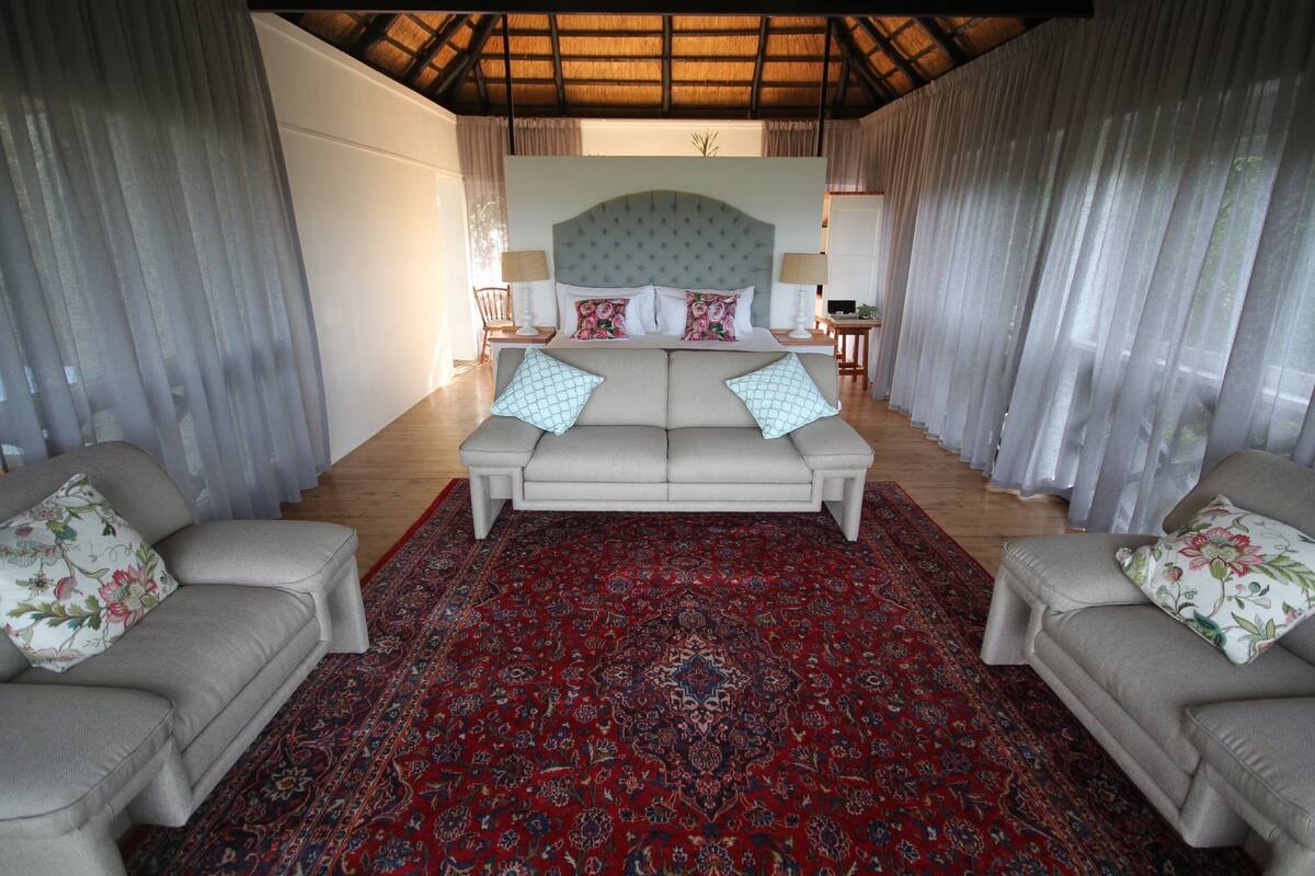 A bedroom with a large bed, two sofas, a red patterned rug, and sheer curtains.