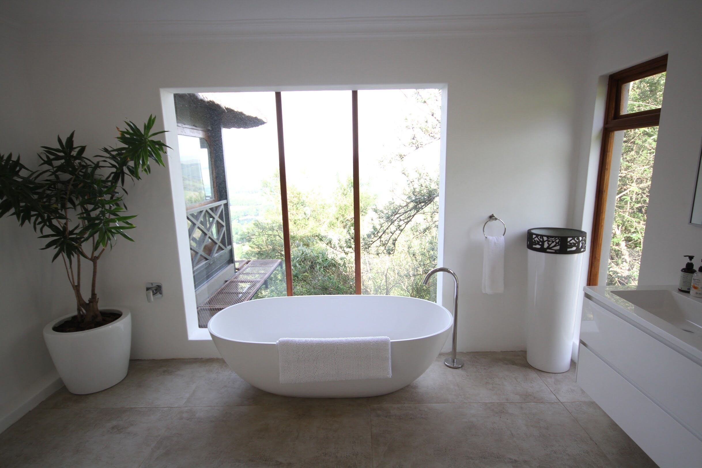 A modern bathroom with a freestanding bathtub, a potted plant, and a view of greenery outside.