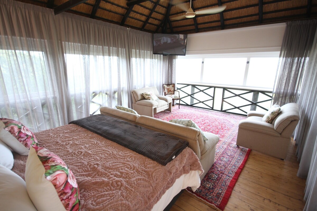 Un dormitorio con una cama grande, dos sillones, un televisor y un balcón con barandilla.