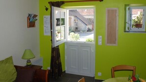 A room with a bright green wall, a wooden door leading outside, a small window with a view of the outside, a table with a chair, and a lamp on a side table.