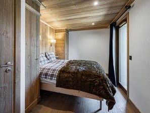 Ein Zimmer mit Holzwänden, einem Bett, einem Nachttisch, einer Wandlampe und einem mit Vorhängen versehenen Fenster.