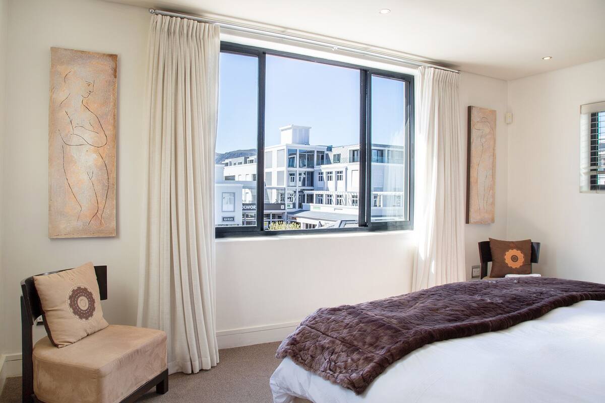 A hotel room with a large window, a bed with a brown blanket, two chairs, and artwork on the walls.