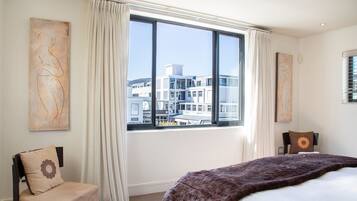 A hotel room with a large window, a bed with a brown blanket, two chairs, and artwork on the walls.