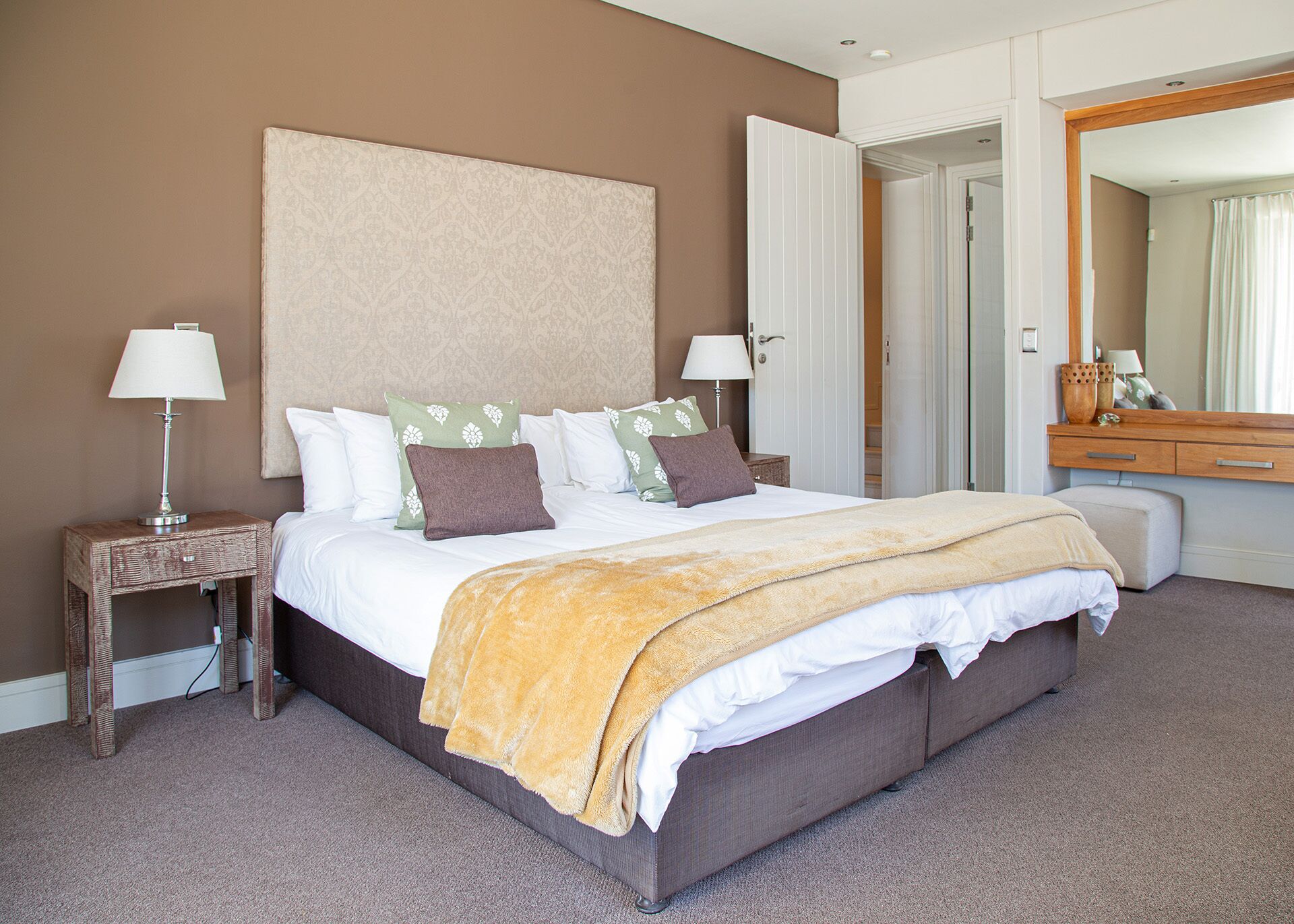 A hotel room with a large bed, two bedside tables with lamps, a wooden nightstand, a mirror, and a door leading to another room.
