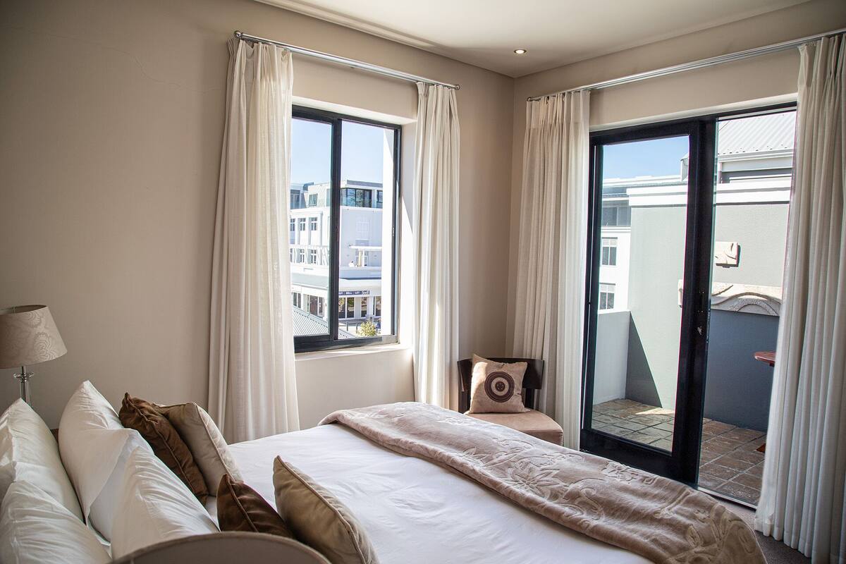 A hotel room with a large bed, a chair, and a view of the city through the windows.