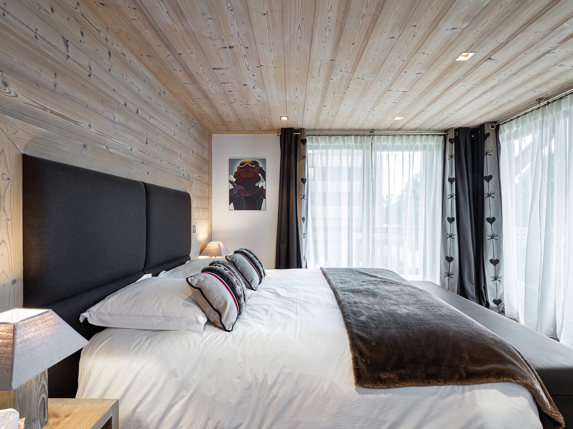 Una camera da letto moderna con soffitto in legno, un letto grande con lenzuola bianche e molti cuscini, un comodino con una lampada e una finestra con tende trasparenti.