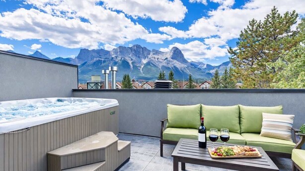 A rooftop terrace with a hot tub, a green sofa, a table with a bottle and glasses, and a scenic mountain view. - Rundleview - Impeccable Design and Private Hot Tub (Canmore)