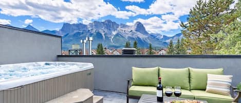 A rooftop terrace with a hot tub, a green sofa, a table with a bottle and glasses, and a scenic mountain view.