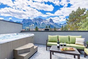 A rooftop terrace with a hot tub, a green sofa, a table with a bottle and glasses, and a scenic mountain view. - Rundleview - Impeccable Design and Private Hot Tub (Canmore)