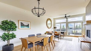 A spacious dining area with a wooden table and chairs, a chandelier, and a large window with a view of mountains. - Rundleview - Impeccable Design and Private Hot Tub (Canmore)