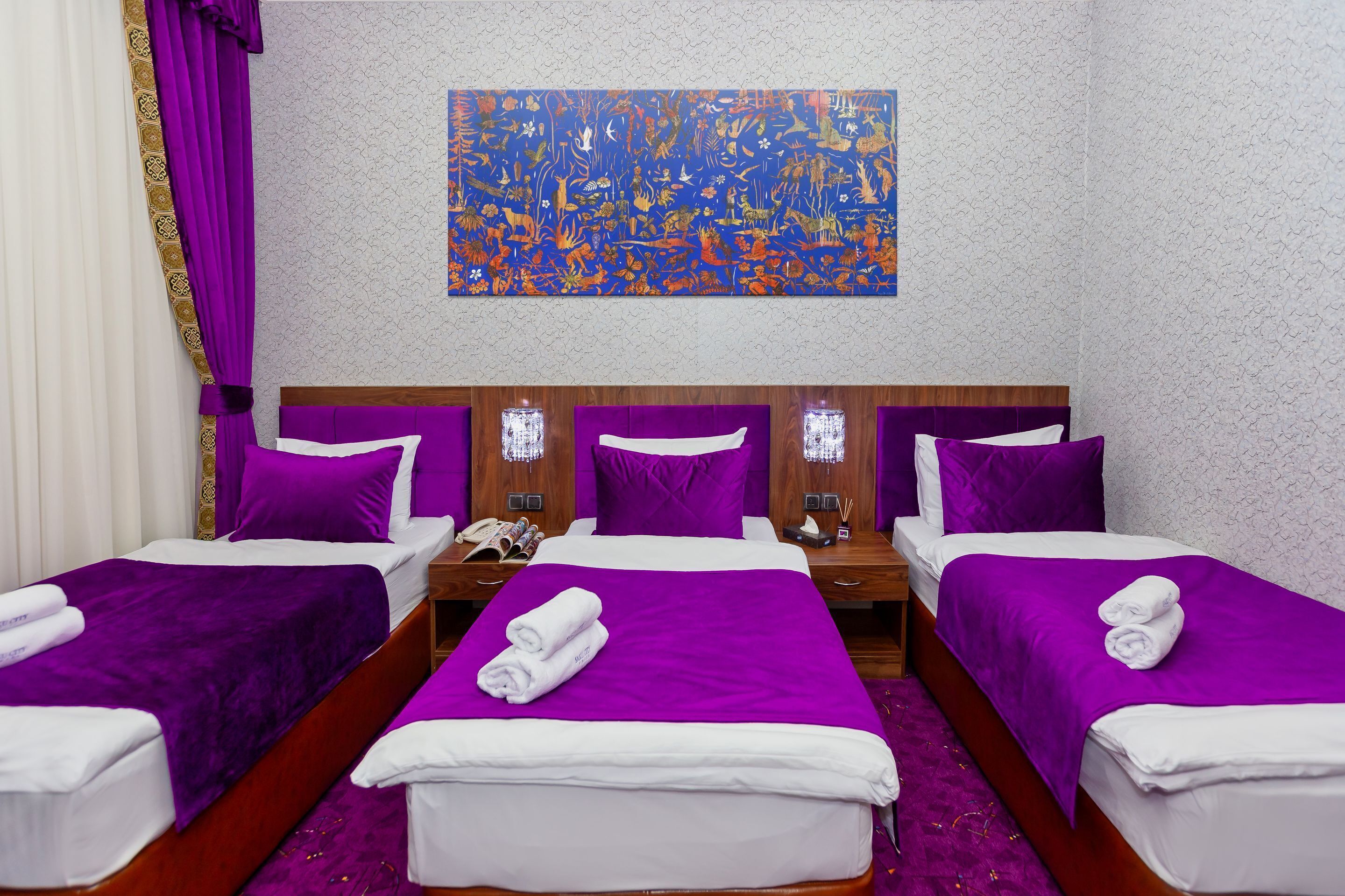 A hotel room with three beds, each with purple bedding and white pillows. There is a large abstract painting on the wall above the beds.