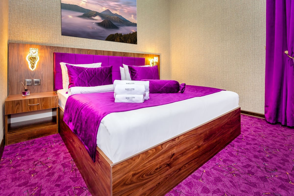 Ein Hotelzimmer mit einem großen Bett, violetten Betttextilien, einem hölzernen Kopfteil, einem Nachttisch, einer Wandleuchte und einem Gemälde einer Berglandschaft.