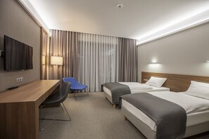Quarto de hotel com escrivaninha, duas camas, televisão, luminária e uma cadeira.