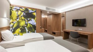 A hotel room with two beds, a desk, a TV, and a large window with a view of trees.