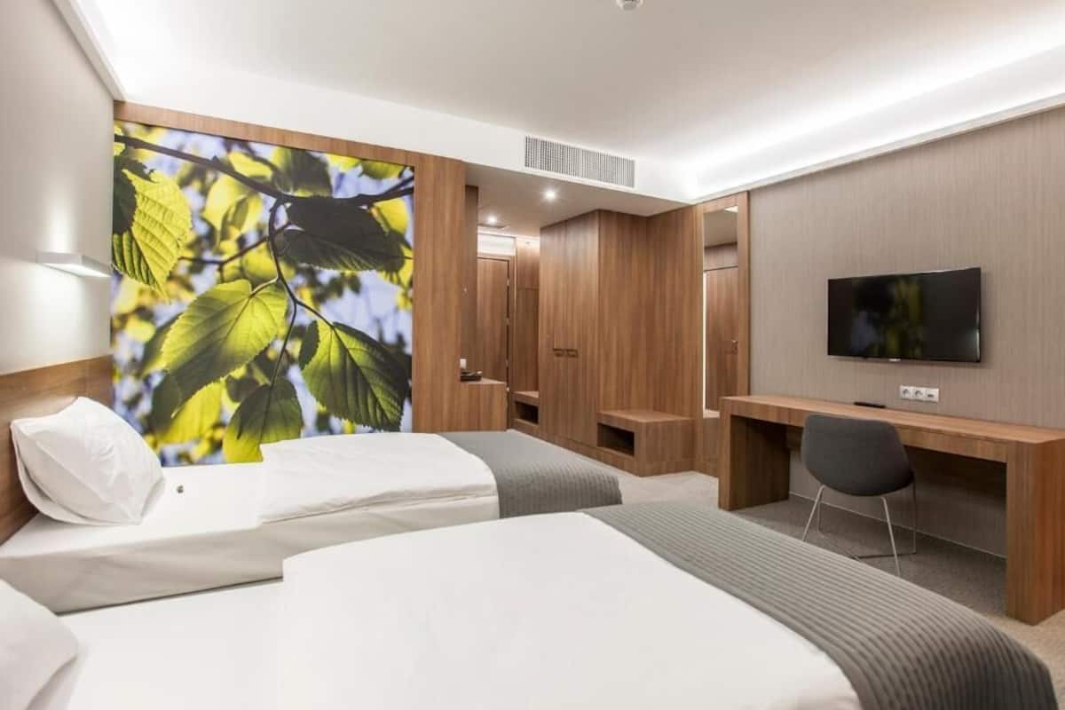 A hotel room with two beds, a desk, a TV, and a large window with a view of trees.