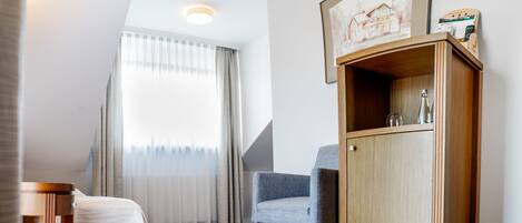 A hotel room with a bed, a wooden cabinet, a chair, and a window with sheer curtains.