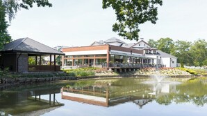 A lakeside hotel with a covered patio, a dining area with outdoor seating, and a reflection in the water.