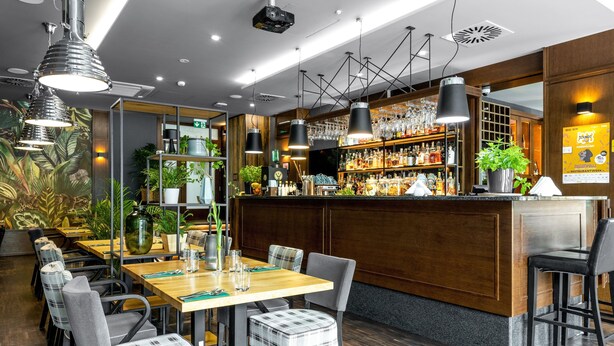 A restaurant with wooden tables and chairs, a bar area with shelves of bottles, and hanging pendant lights.