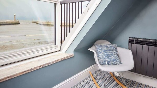 A corner room with a view of the sea, a white chair with a patterned cushion, a striped rug, and a window with a wooden frame.