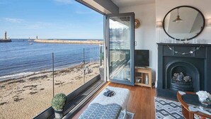 A room with a large window overlooking a beach and the sea, a fireplace, and a seating area with a rug.