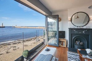 A room with a large window overlooking a beach and the sea, a fireplace, and a seating area with a rug. - Host & Stay | No 3 Sandside (Whitby)