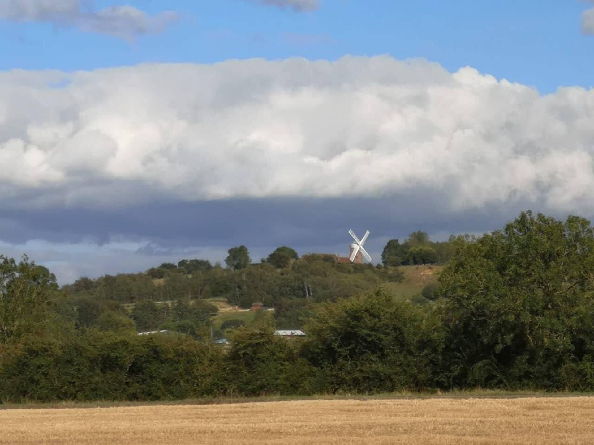 A windmill on a hill with a field in the foreground and a cloudy sky.