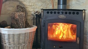 A wood-burning stove with a visible fire, a stone wall, a woven basket, and a hat on the wall. - Quiet vacation rental for 5 people with a safe swimming pool (Salviac)