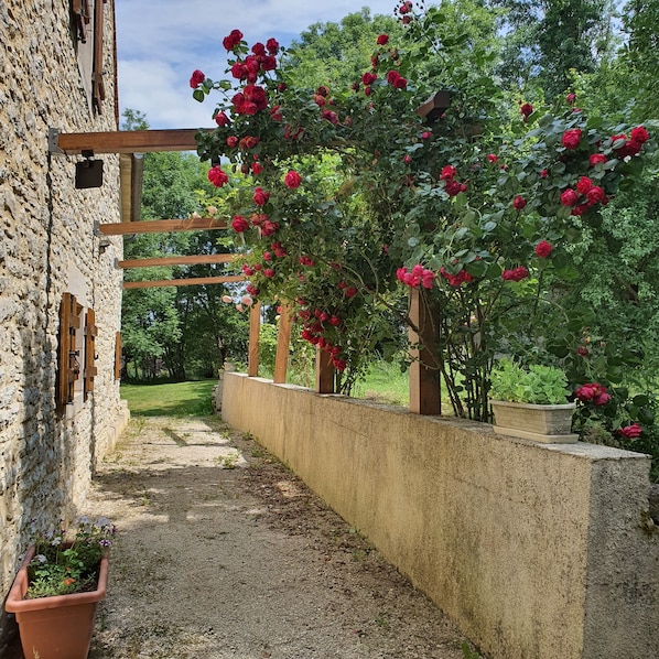 Un bâtiment en pierre surmonté d’une pergola en bois recouverte de roses rouges.