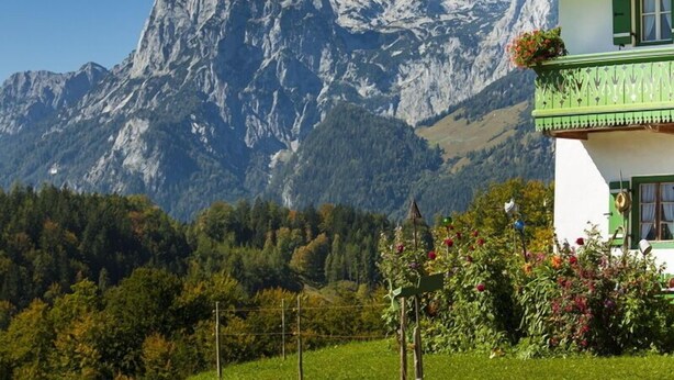Una casa con montañas al fondo, un balcón lleno de flores y un cielo despejado.