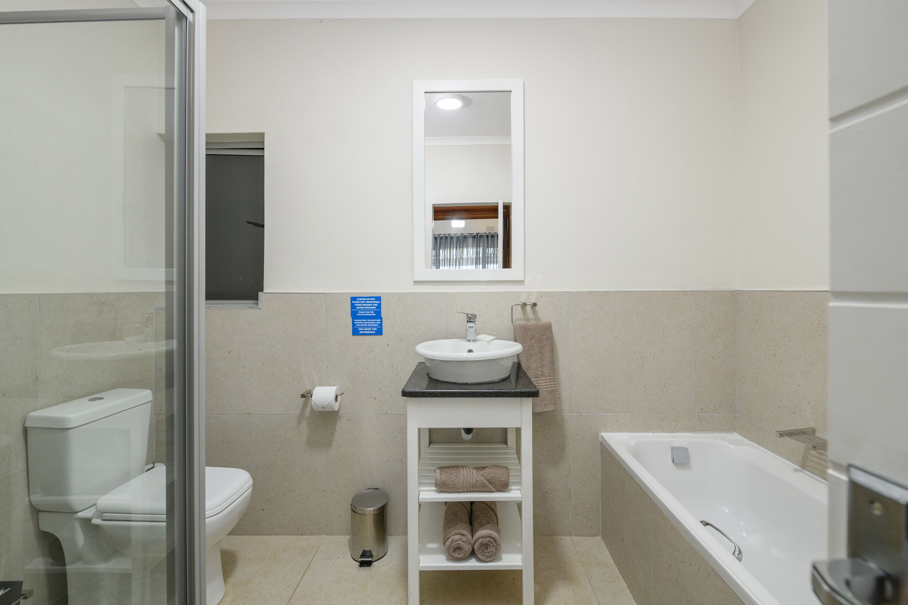 Een badkamer met toilet, wastafel en bad. Er hangt een spiegel boven de wastafel en rechts bevindt zich een handdoekhouder.