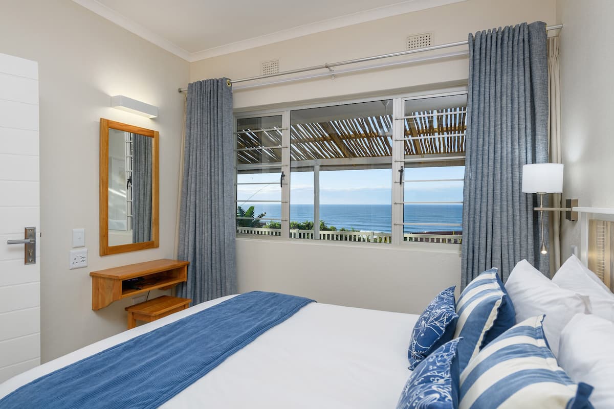 A bedroom with a bed, a nightstand, a mirror, a window with curtains, and a view of the sea.