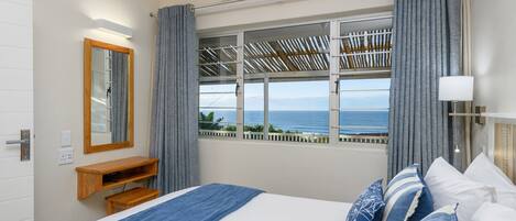 Un dormitorio con cama, mesita de noche, espejo, ventana con cortinas y vista al mar.