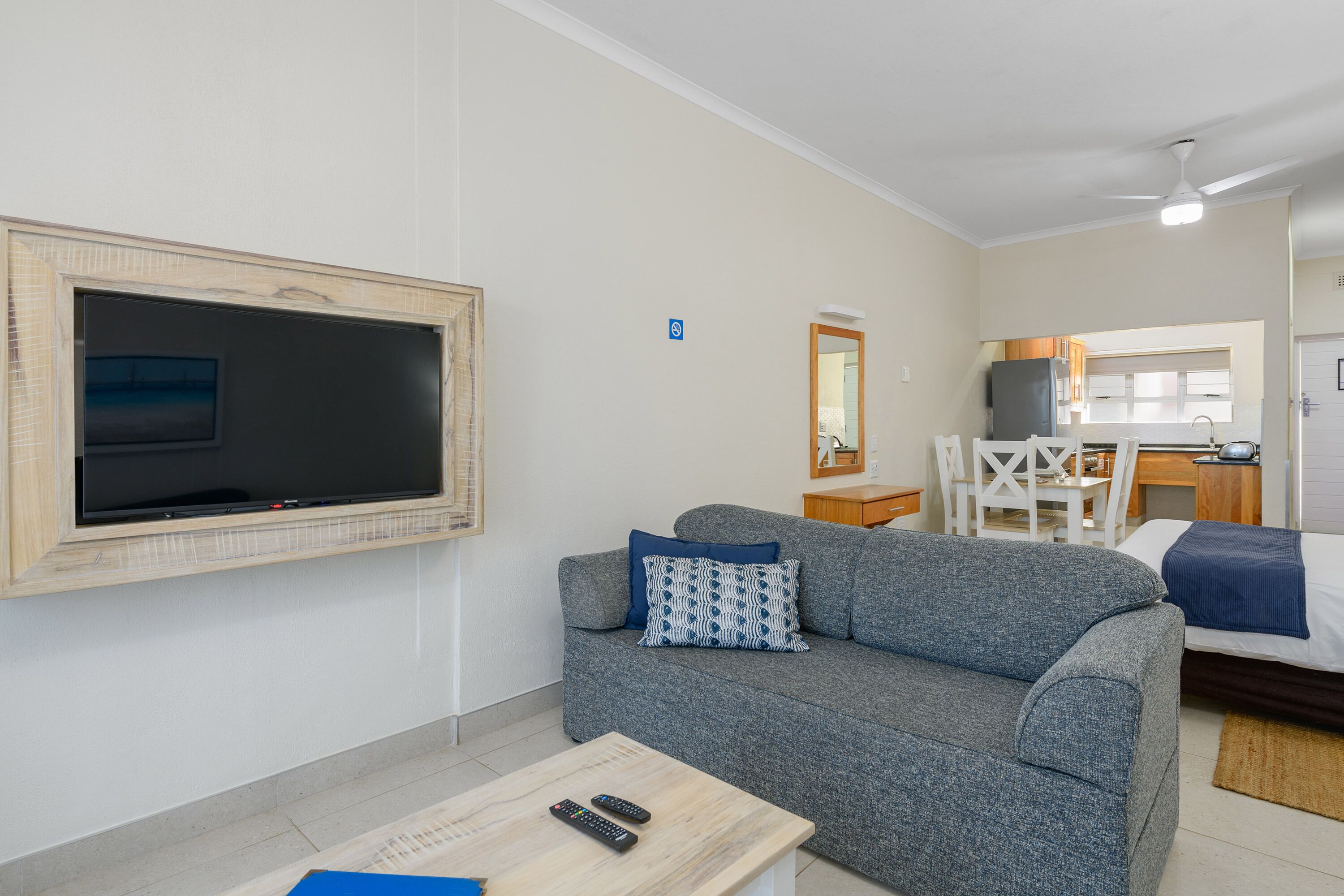 A modern living room with a grey sofa, a flat-screen TV mounted on the wall, a small wooden coffee table, and a dining area with a white table and chairs.