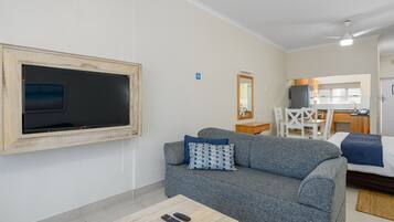 A modern living room with a grey sofa, a flat-screen TV mounted on the wall, a small wooden coffee table, and a dining area with a white table and chairs.