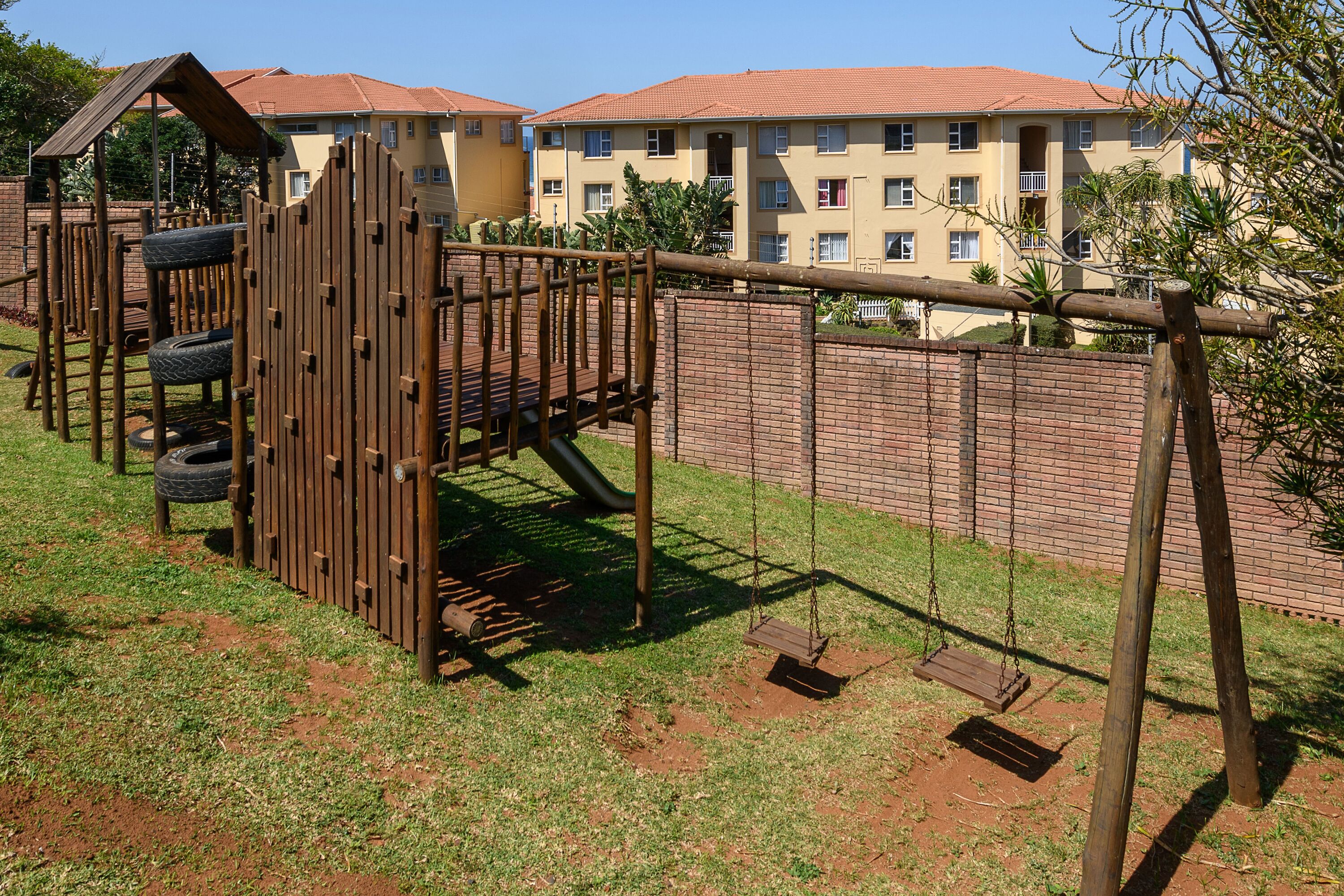 A playground with a wooden climbing structure and swings, surrounded by a brick wall and a building with a red roof in the background.