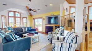 A spacious living room with a large TV, comfortable seating, a fireplace, and a staircase. - Solstice by the Sea | 7BR Carova Beach 4x4 OBX | Pool, Sauna & Theater (Corolla)