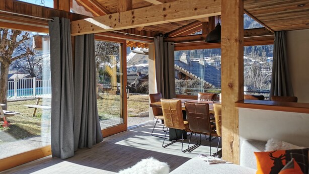 Una habitación espaciosa con vigas de madera, un comedor con sillas y ventanas amplias que ofrecen vistas de un paisaje montañoso.