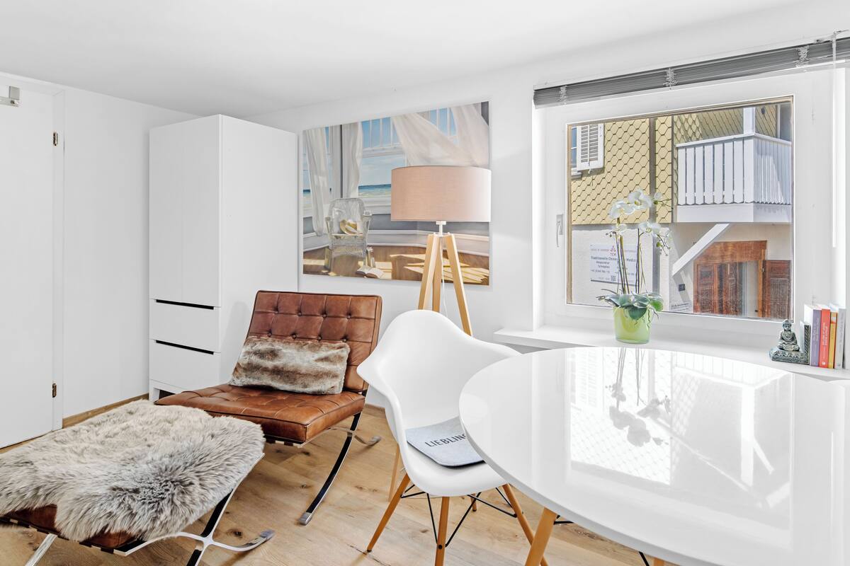 A modern living room with a white sofa, a brown leather armchair, a white dining table, and a large window with a view of a building.