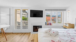 A modern hotel room with a fireplace, a flat-screen TV, a balcony with a view of buildings and trees, and a neatly made bed with towels. - HouseofHarmony (Stafa)