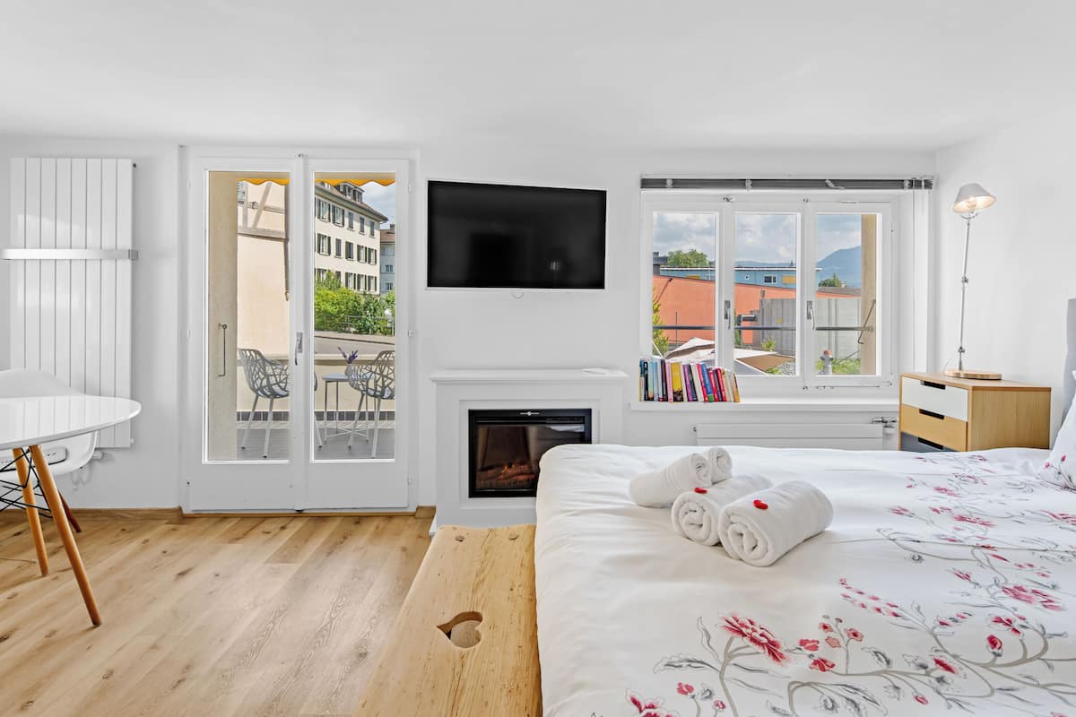 A modern hotel room with a fireplace, a flat-screen TV, a balcony with a view of buildings and trees, and a neatly made bed with towels.