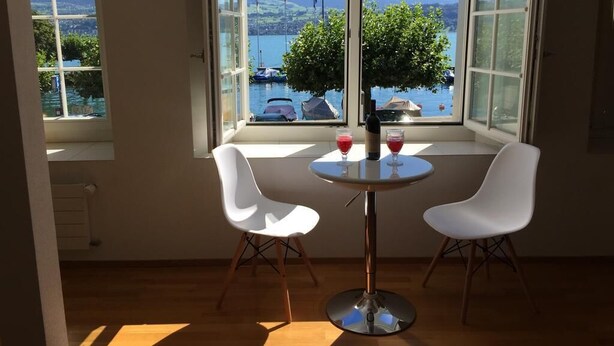 A room with a large window overlooking a body of water and boats, a round table with two white chairs, and a small tree in the window. - HouseofHarmony (Stafa)