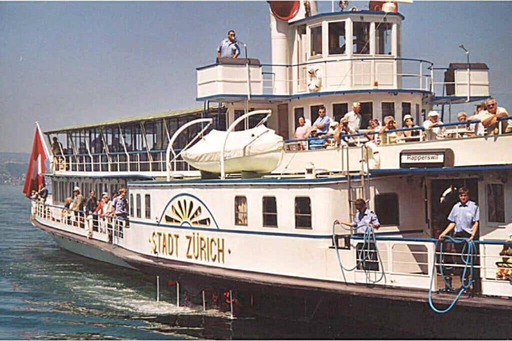 Un bateau portant le nom « Stadt Zürich » sur sa coque, des passagers sur le pont et une personne debout sur le pont supérieur.